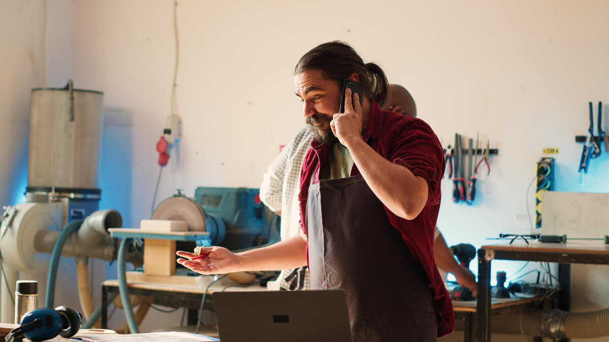 A workshop owner taking a customer call at his bench, laptop open beside him