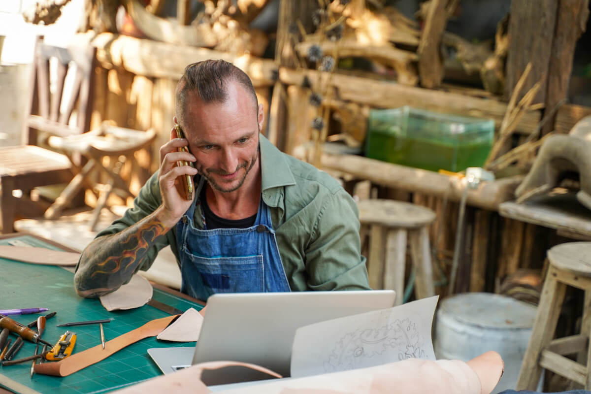 Leather-goods maker on the phone at his workbench, laptop open, mid-conversation