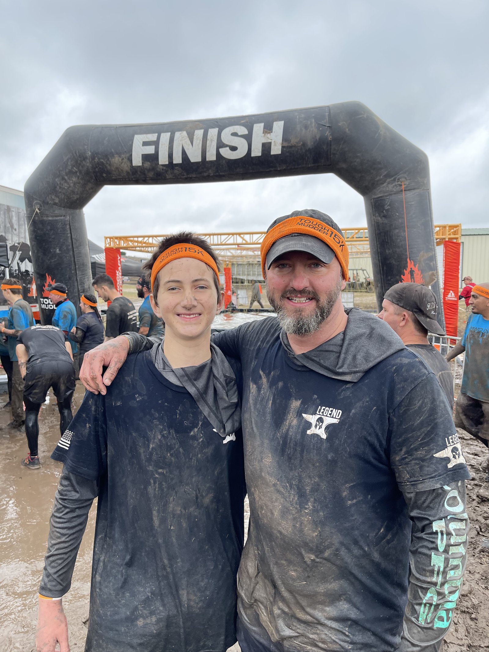 Shawn Hurlburt and his son, muddy and smiling under the finish arch at a Tough Mudder race