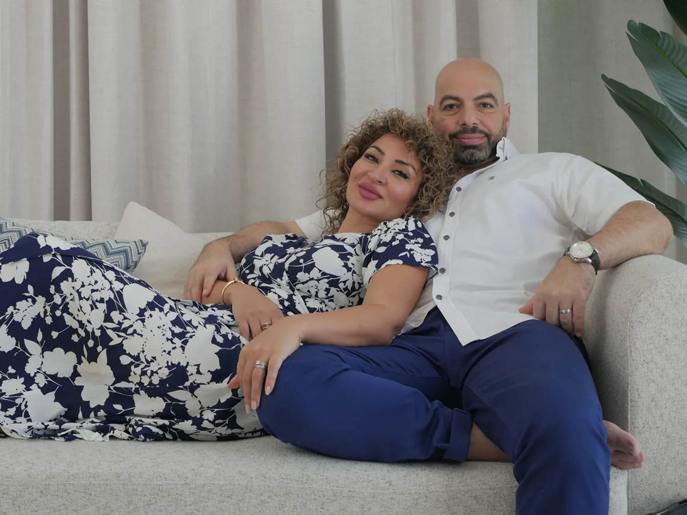 Fatima resting against Ramy on a light grey sofa, both dressed in matching navy and white, relaxed and at ease together.