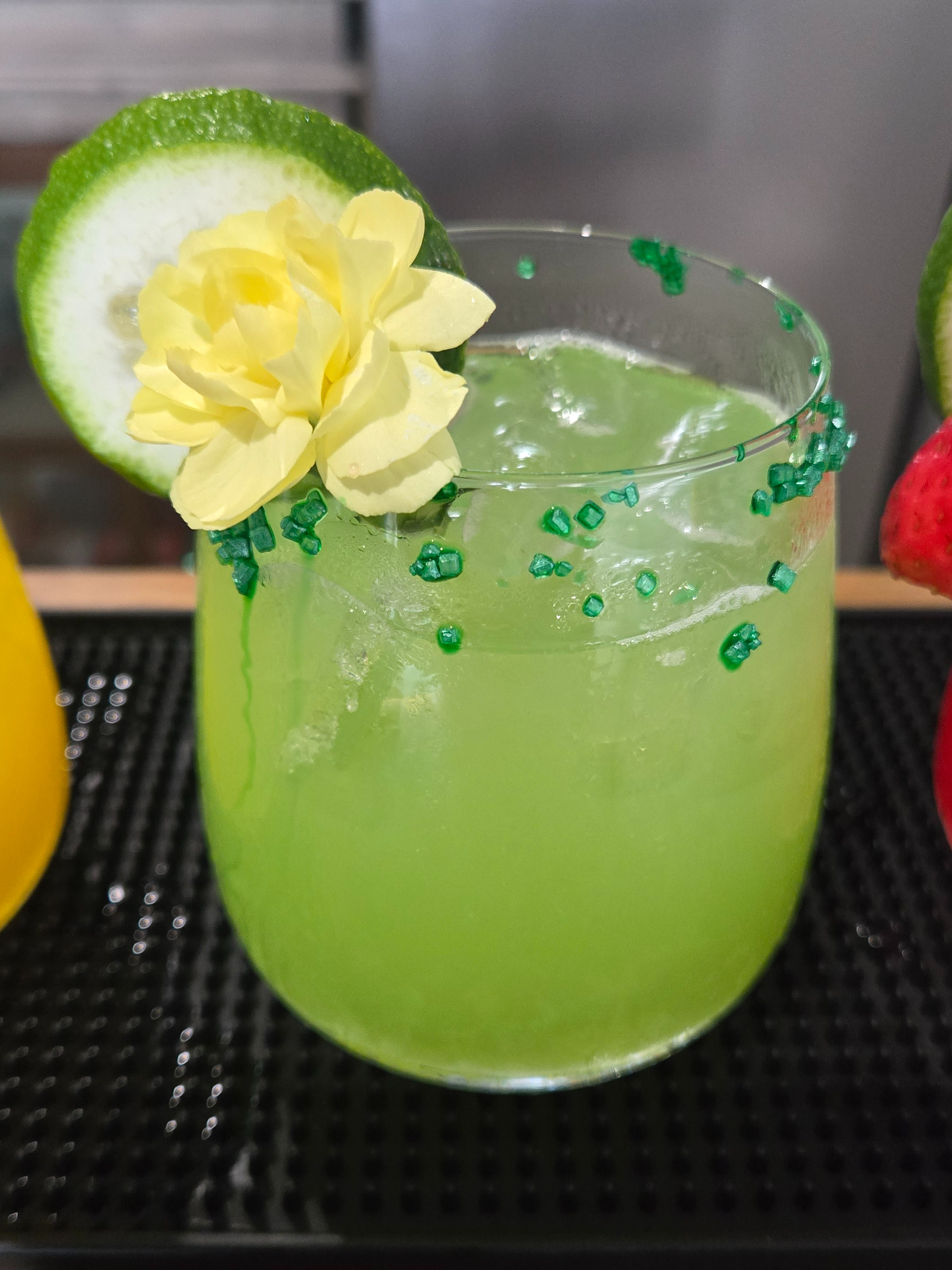 Green cocktail garnished with a cucumber wheel, yellow flower, and green sugar rim on a bar mat