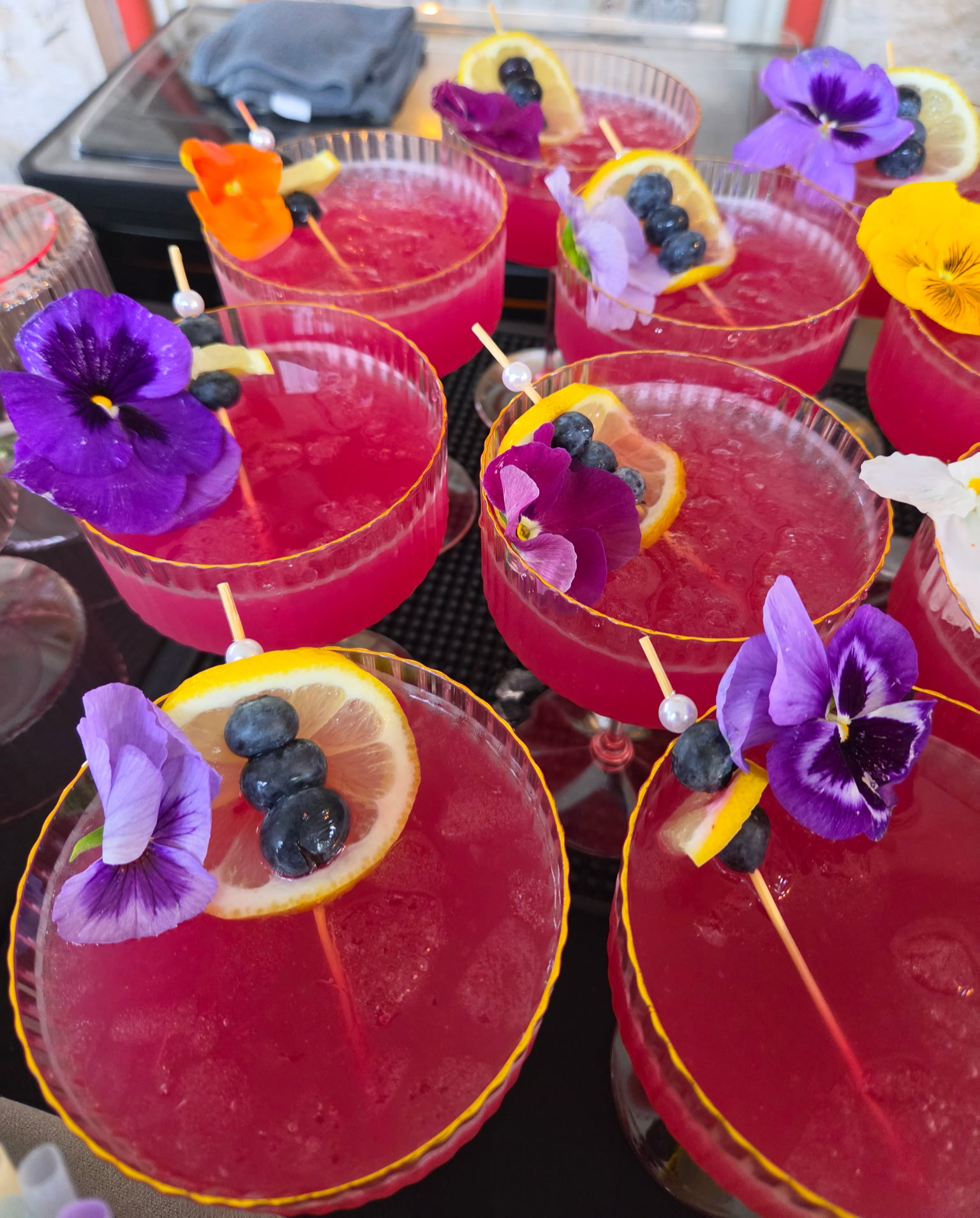 Rows of magenta cocktails in gold-rimmed coupe glasses garnished with lemon wheels, blueberries, and edible pansies