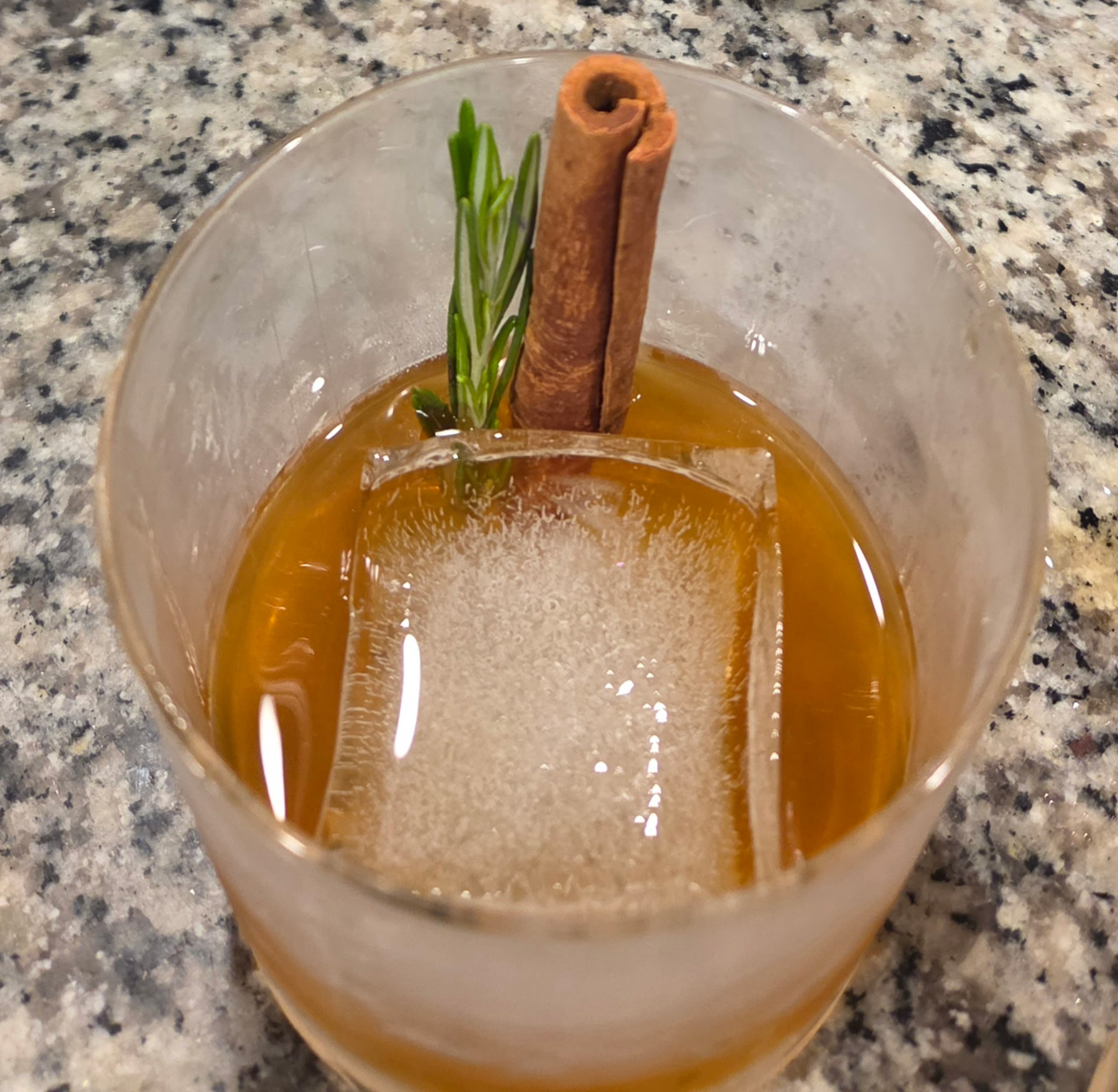 Whiskey cocktail with a large ice cube, rosemary sprig, and cinnamon stick garnish