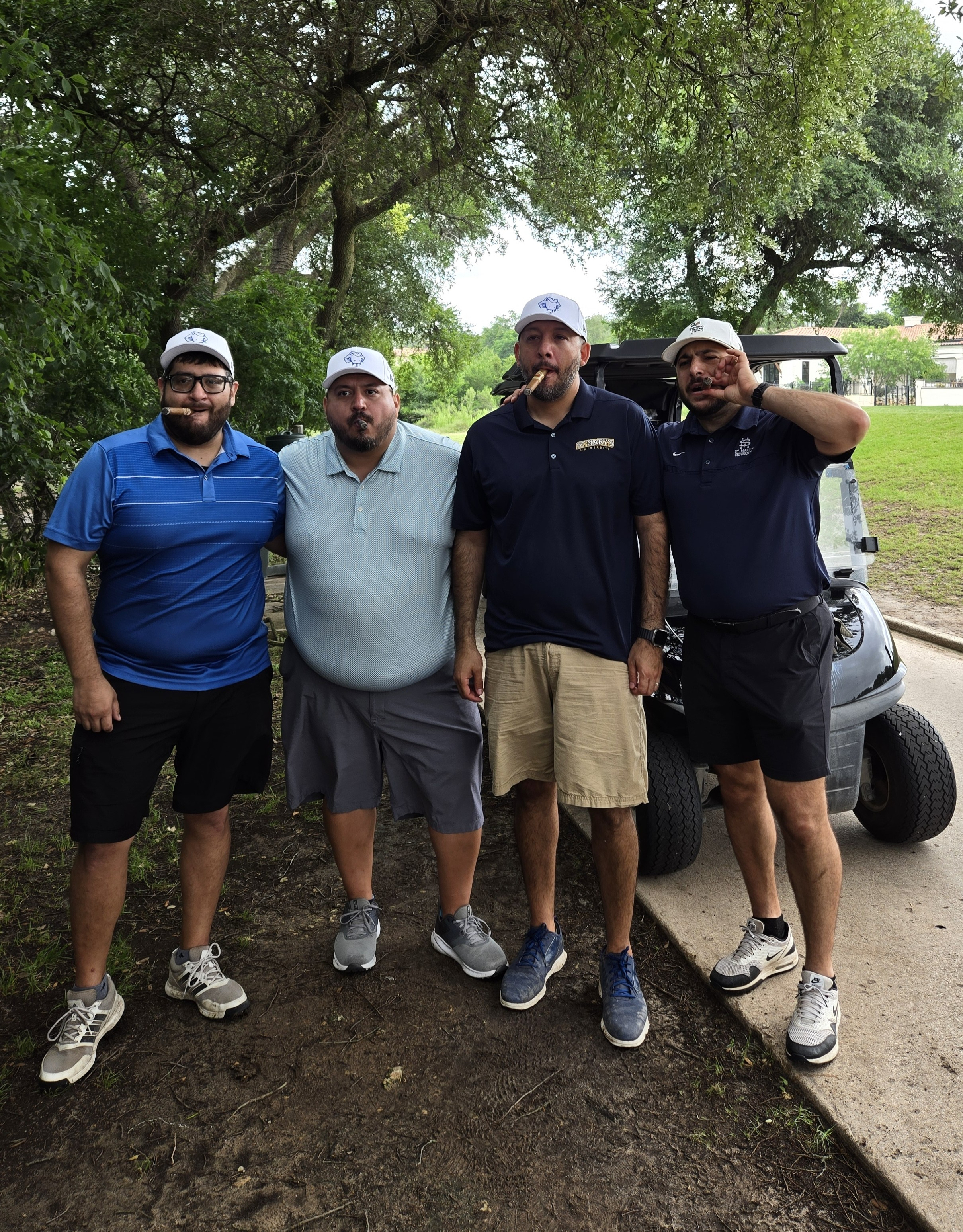 Four golfers enjoying cigars beside a golf cart under oak trees at a tournament