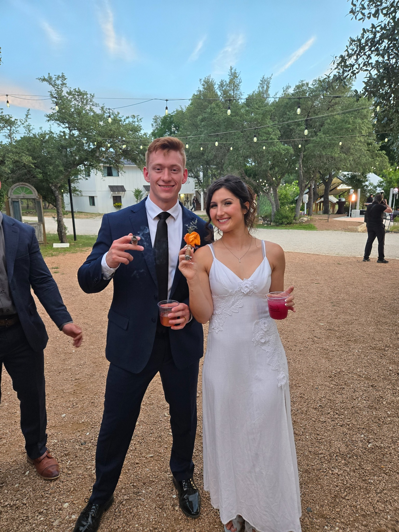 Bride and groom holding cigars and cocktails under string lights at their wedding