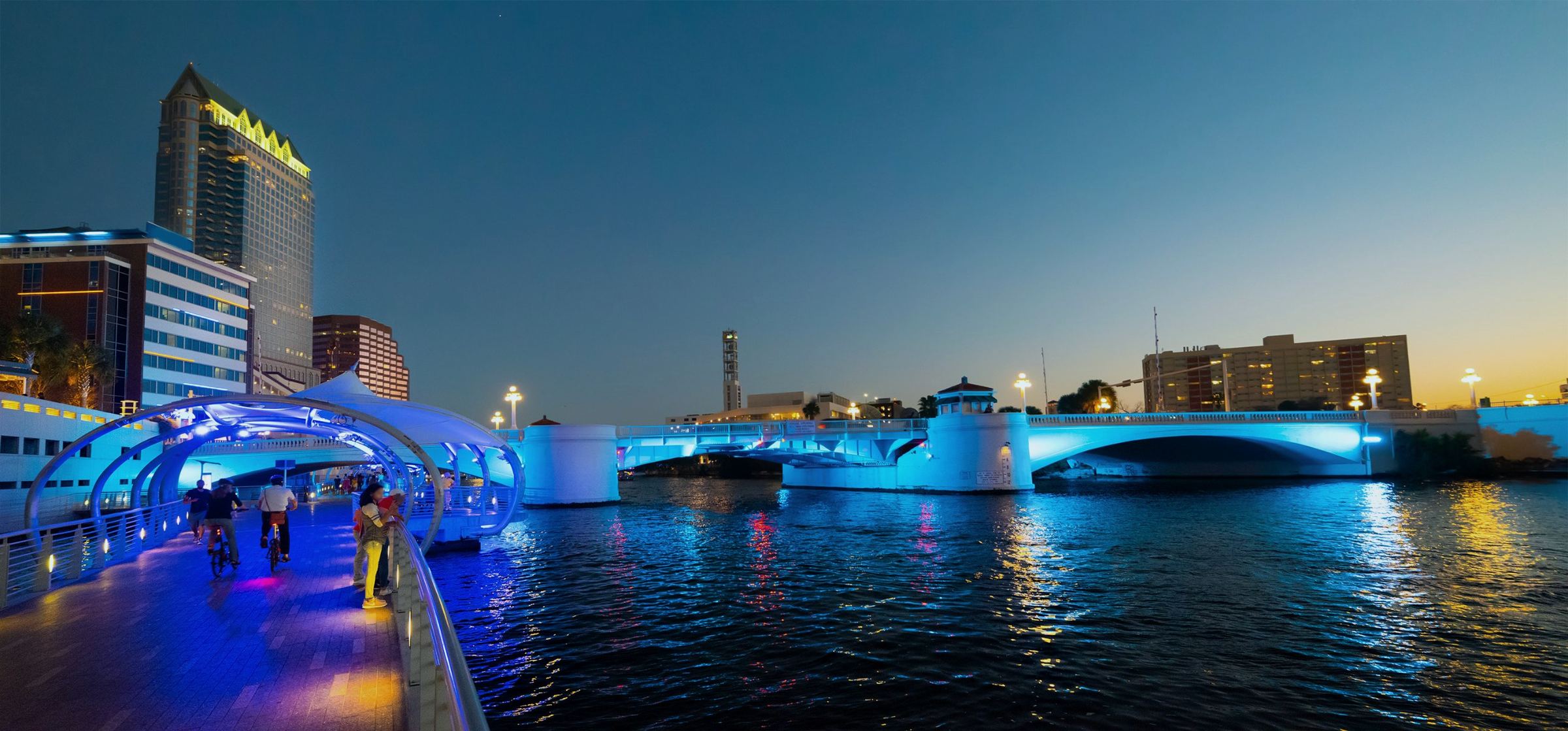 The Tampa Riverwalk at dusk, with the lit bridge over the Hillsborough River