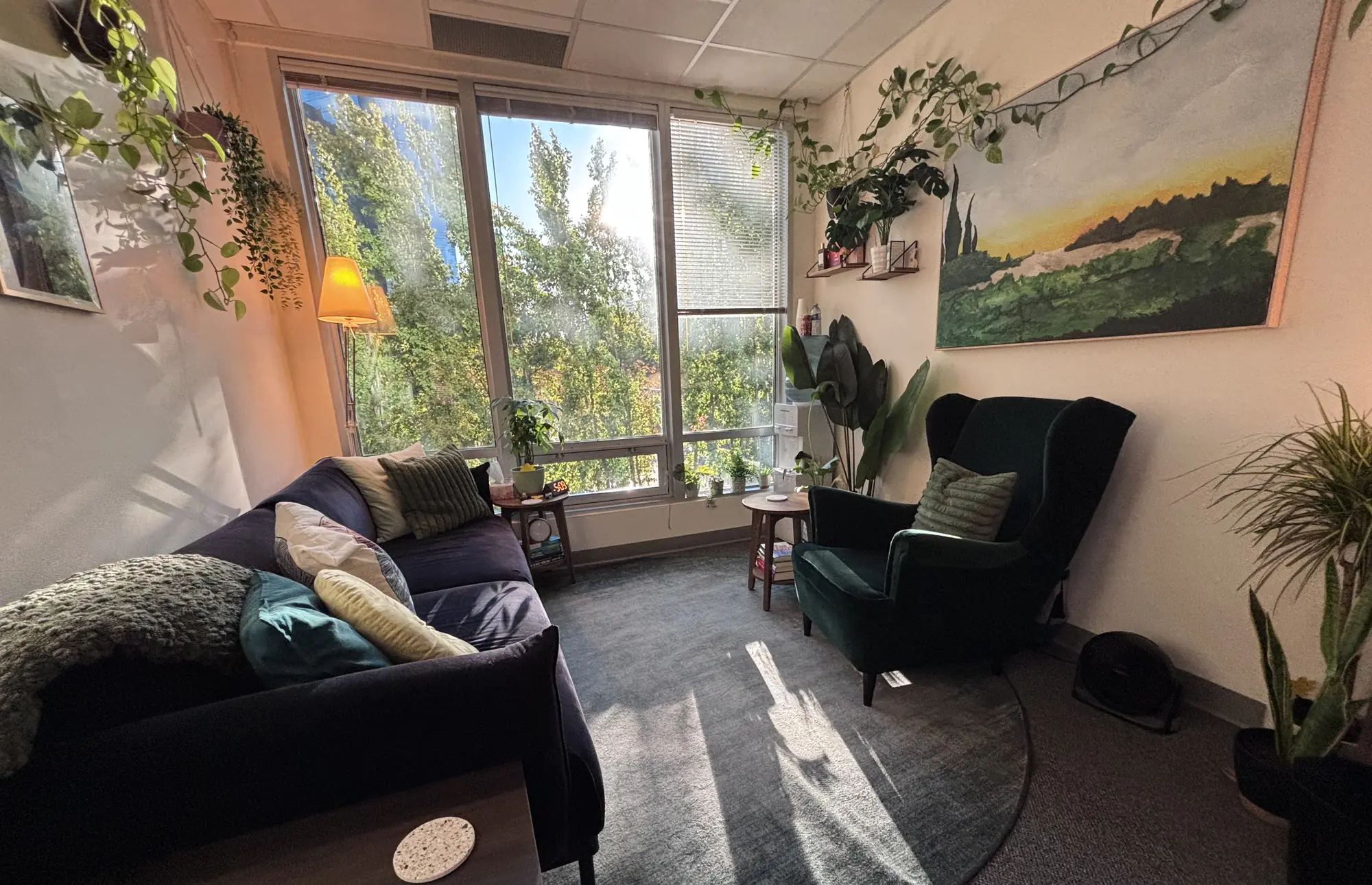 Wide view of a calm counselling office with a navy sofa, green armchair, round rug and tall windows looking onto trees