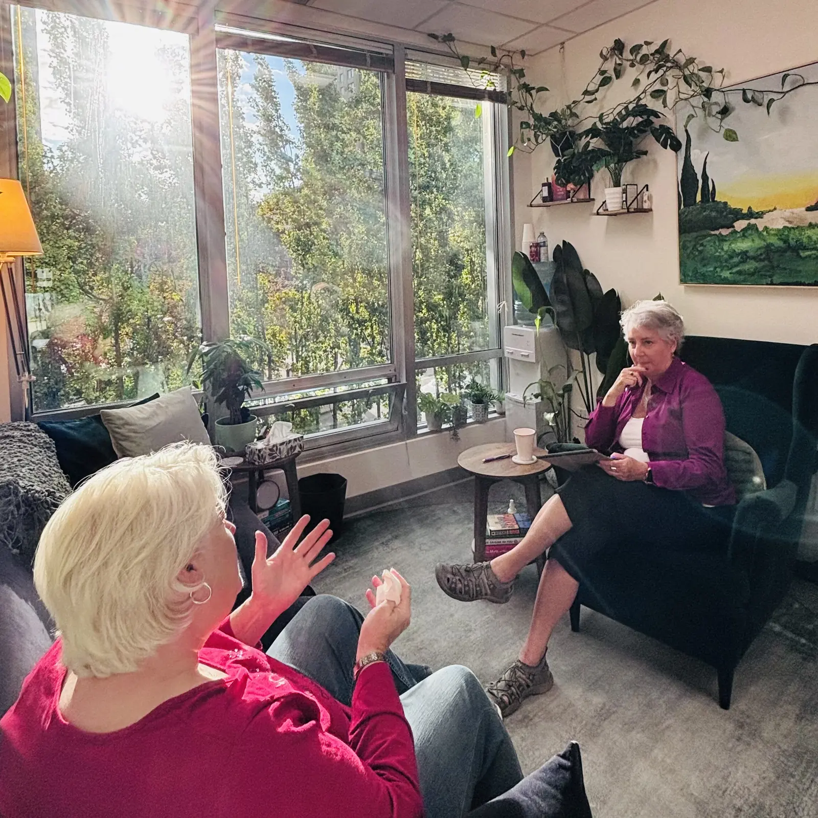 Melinda Freitas smiling in a green armchair with a tablet while a client with white hair sits across from her in her bright, plant-filled office