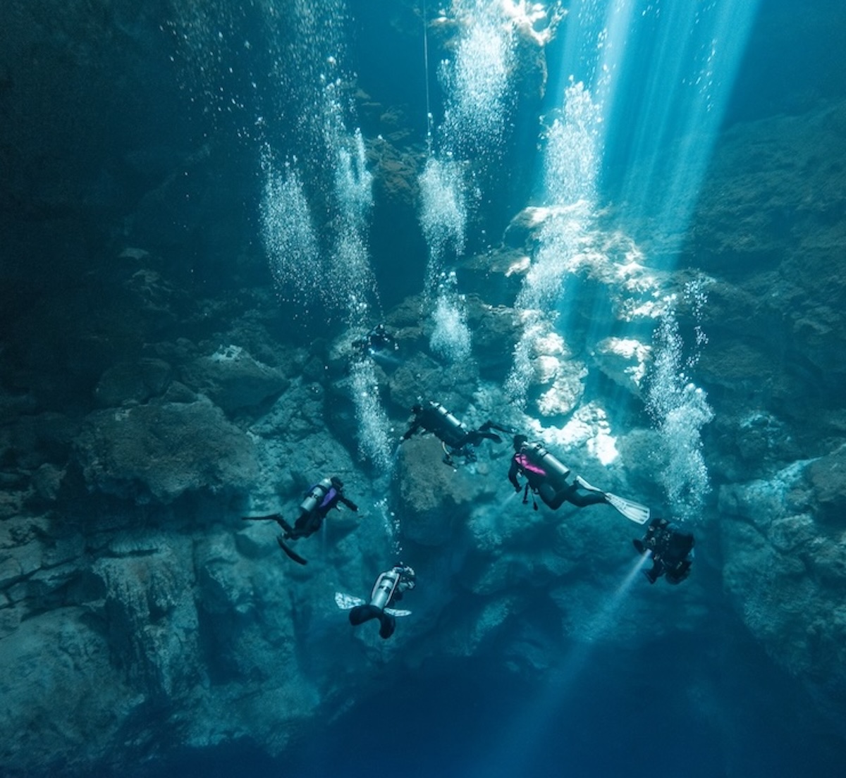 scuba divers surrounded by light beams descending into El Pit cenote cavern in Tulum, Mexico