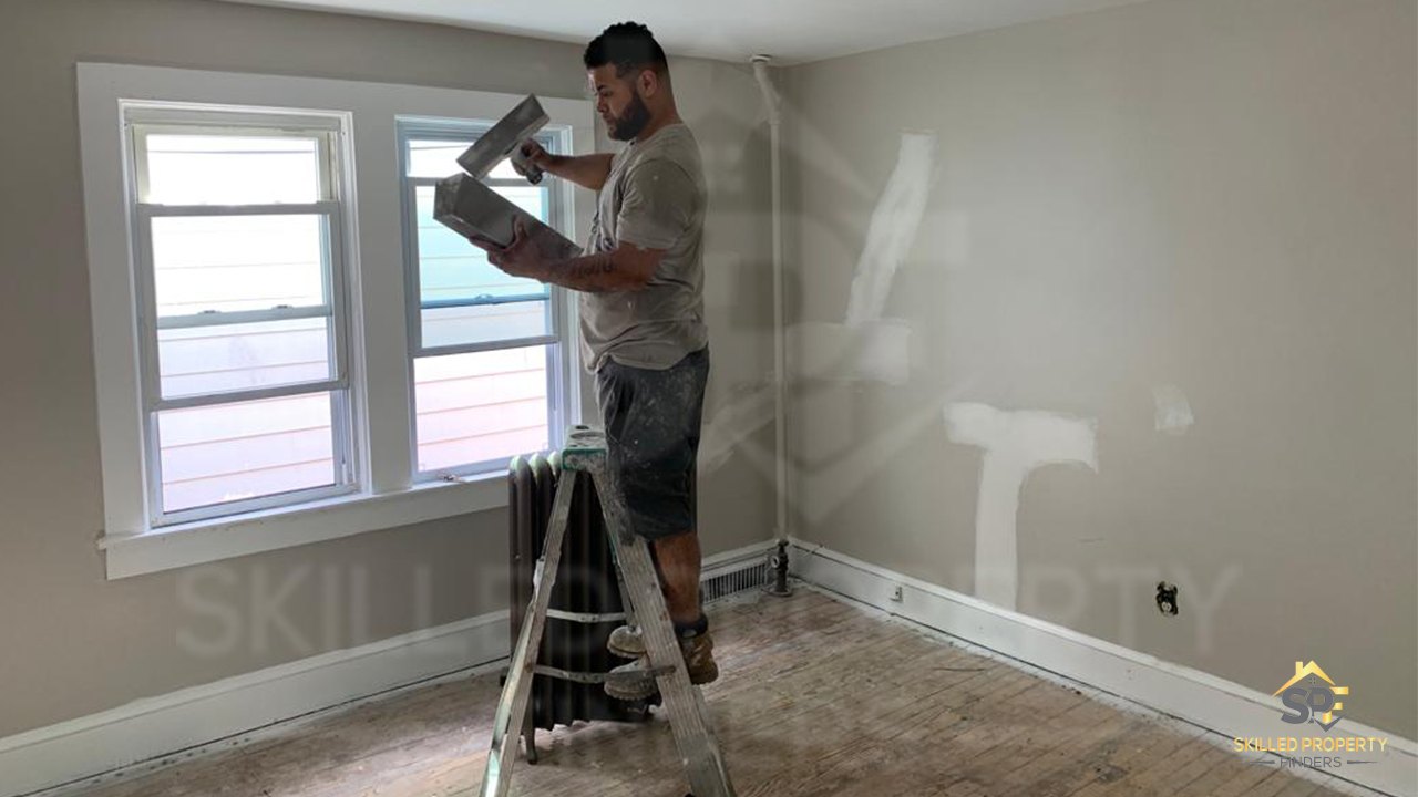 Partway through the rebuild at Empire Street: a tradesman on a ladder skim-coating a bedroom wall, subfloor exposed and the old radiator still in place.