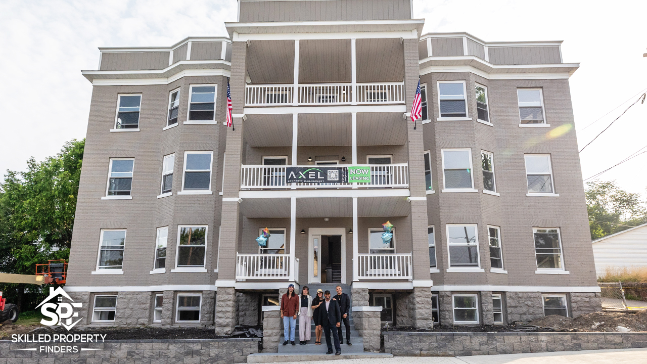 The renovated 12-unit apartment building on Vine Street, Scranton, from the front on opening day: grey brick with white stacked porches, two American flags, a Now Leasing banner, and four people standing on the front steps.