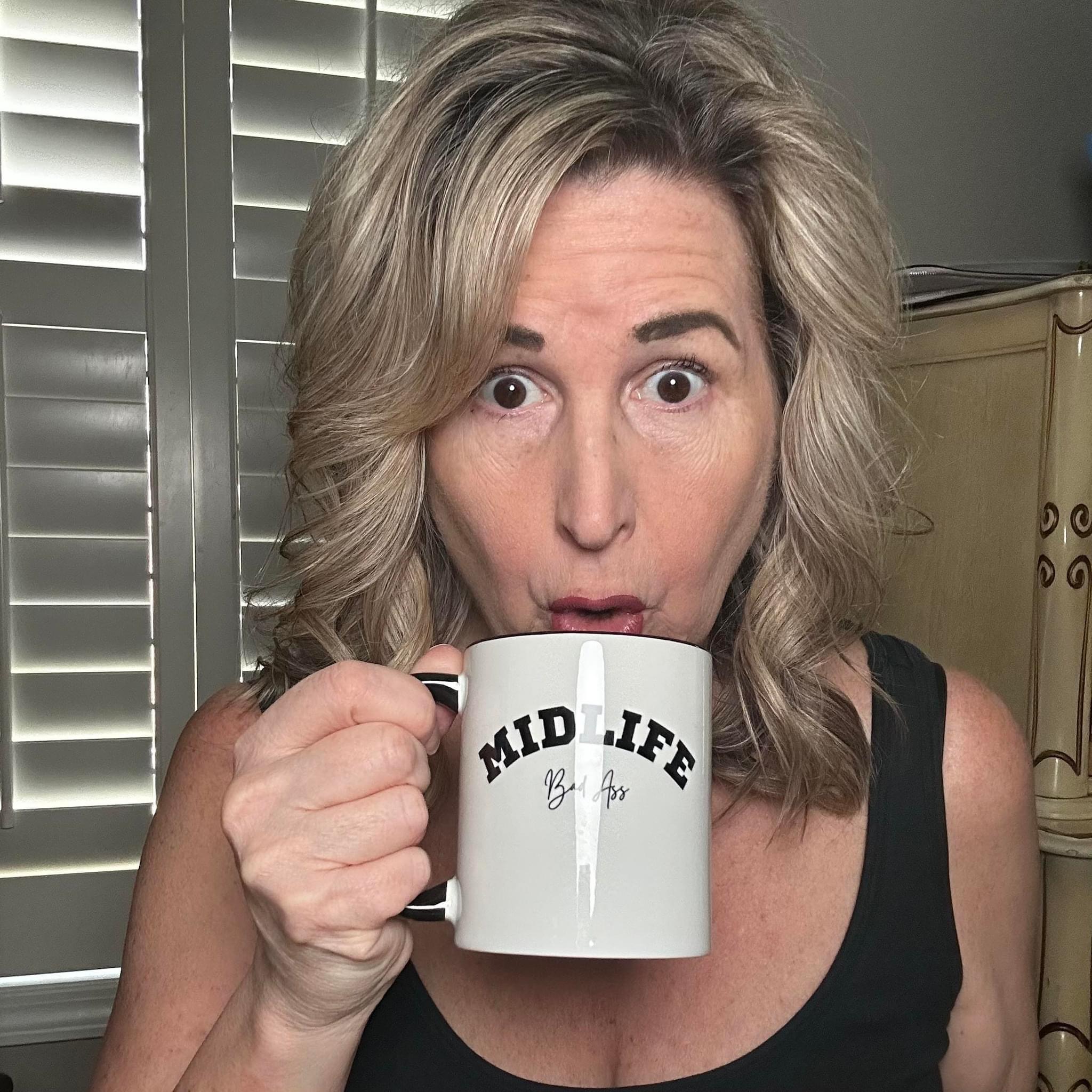 A woman with shoulder-length wavy blonde hair wearing a black tank top holds a white coffee mug with 'MIDLiFE BadAss' printed on it. She has a surprised expression while sipping from the mug. The background features window shutters and a wooden dresser.