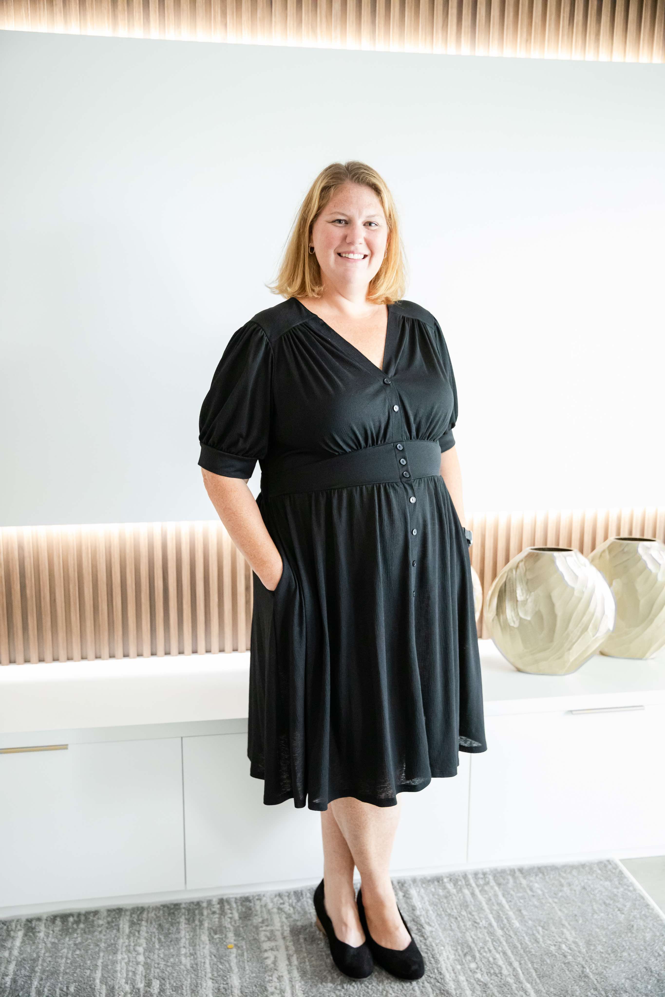 A smiling woman with shoulder-length blonde hair wearing a black button-down dress and black flats. She stands with her hands in her pockets against a modern, well-lit background featuring decorative vases and a textured wall panel.