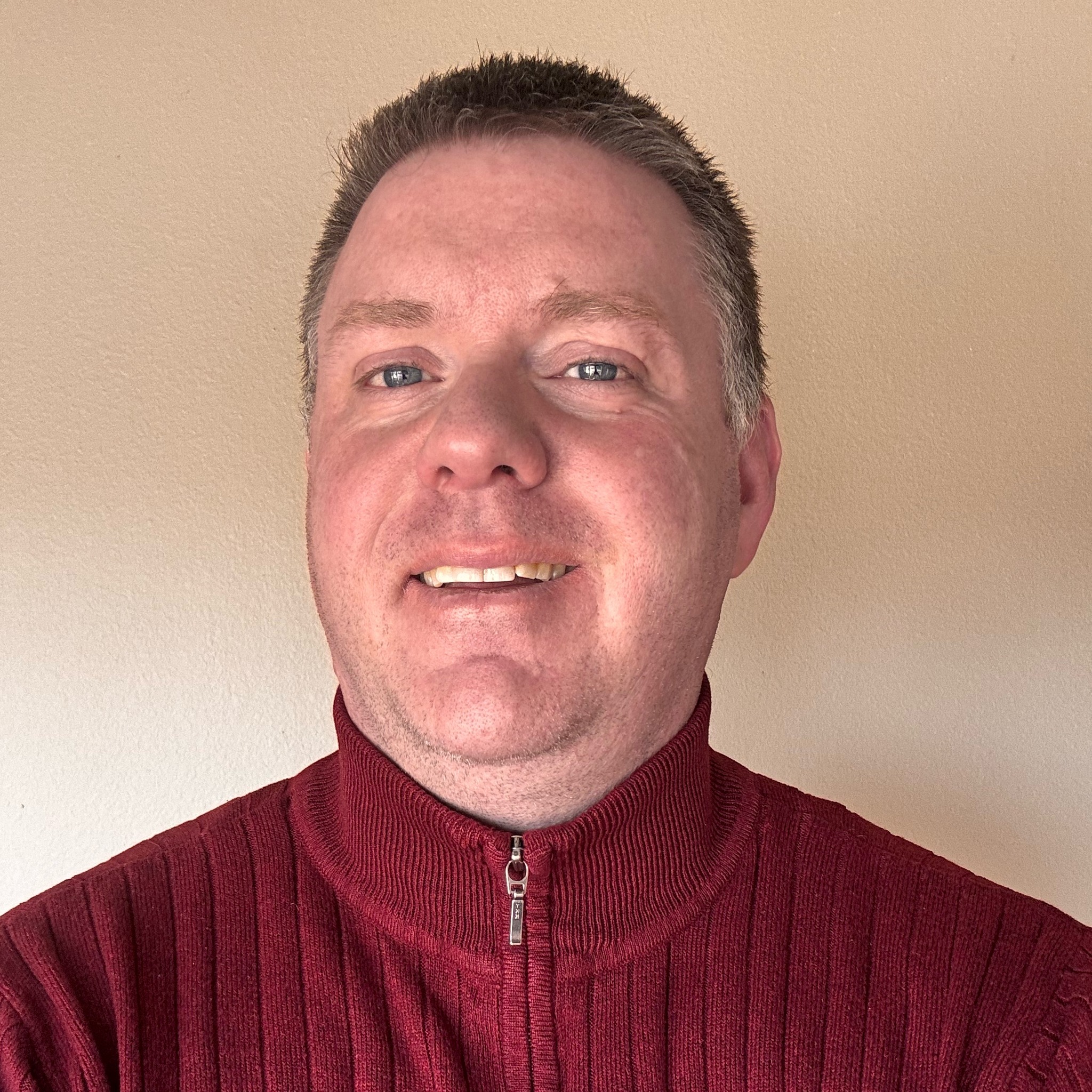 A smiling man with short-cropped hair wearing a red ribbed zip-up sweater. He stands against a plain beige background.