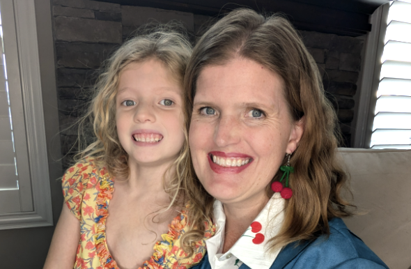 A smiling woman with shoulder-length brown hair wearing cherry-themed earrings and a blue jacket with a white collared shirt, posing with a young girl who has long curly blonde hair and is wearing a colorful floral dress. They are sitting together in a warmly lit room with a stone wall and window shutters in the background.