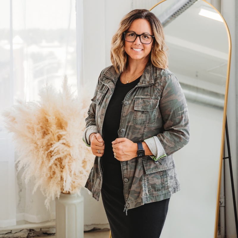 Woman in a camo jacket standing indoors near a floor mirror and pampas grass, smiling with hands holding her jacket.