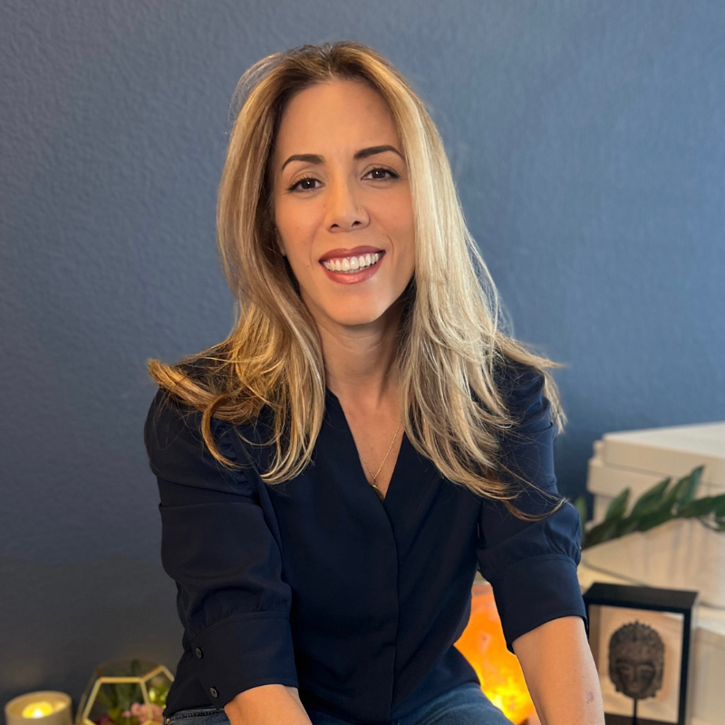 Woman sitting in front of a navy blue wall, smiling warmly, with soft lighting and candles in the background.