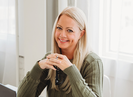 Professional woman with long blonde hair smiling at her desk, hands folded and confident expression.
