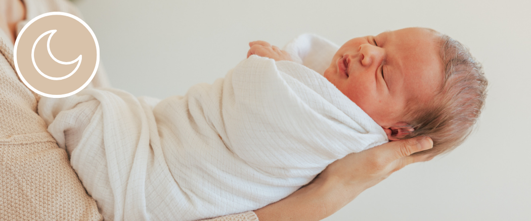 Sleeping newborn baby wrapped in a white swaddle and supported in an adult's arms.