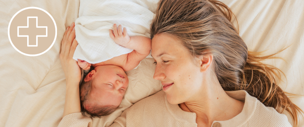 Mother smiling at her newborn baby while lying together on a bed.