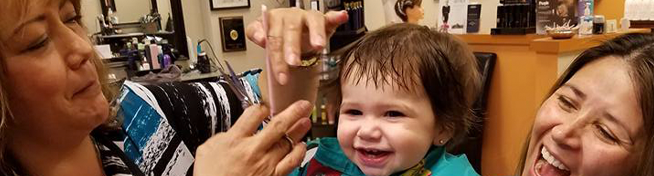 Sonia giving a smiling toddler a haircut while a parent laughs beside them