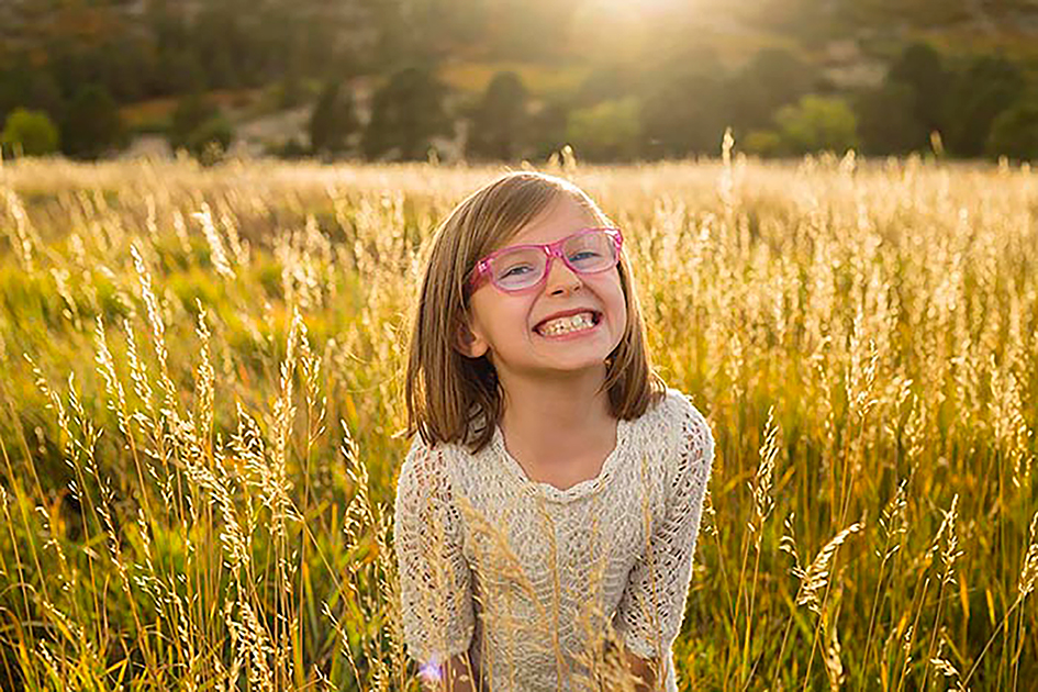 Golden hour outdoor portrait of a smiling young girl in a lace dress with pink glasses, standing in a sunlit field of tall grass, natural light child photography in Colorado Springs with warm, candid emotion.