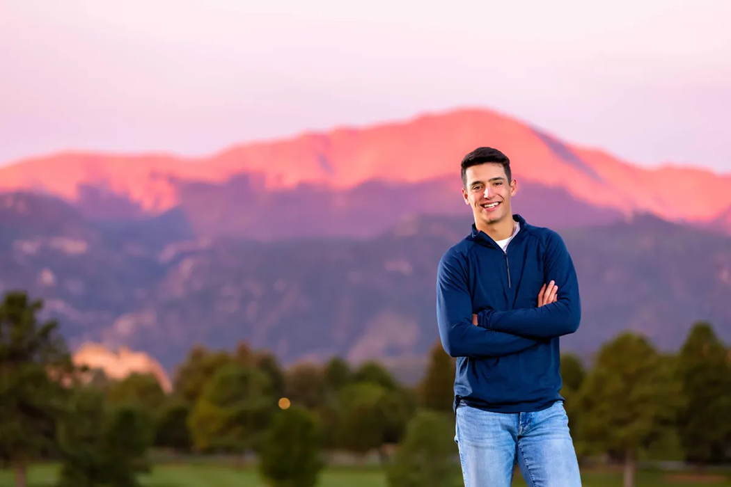 Sunset senior portrait of a smiling young man in casual clothing standing in front of scenic mountain landscape, natural light outdoor photography in Colorado Springs with warm, golden tones and vibrant background.