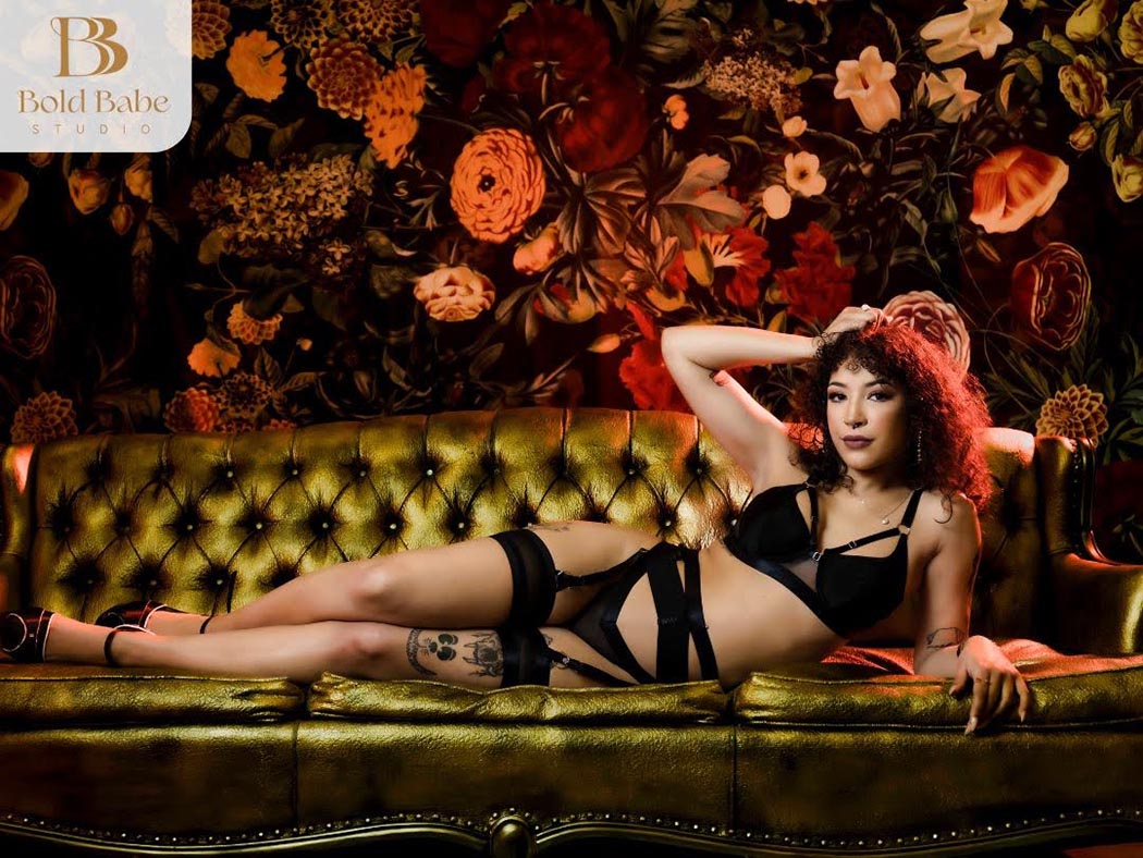 Empowering boudoir portrait of confident woman in black lingerie reclining on a vintage gold velvet sofa with dramatic floral backdrop, luxury studio photography in Colorado Springs by Bold Babe Studio.