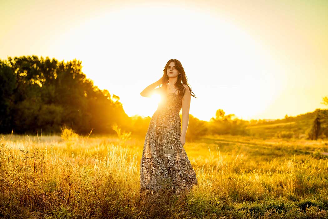 Golden hour portrait of a young woman in a flowing patterned dress standing in a sunlit field, dreamy outdoor photography in Colorado Springs with warm backlighting and scenic countryside setting.