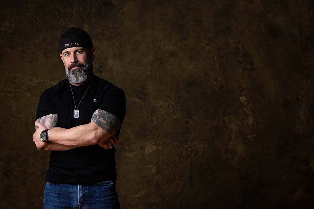 Moody studio portrait of muscular bearded man with tattoos and black t-shirt posing confidently against a dark textured backdrop, edgy personal branding photography in Colorado Springs with strong masculine style.