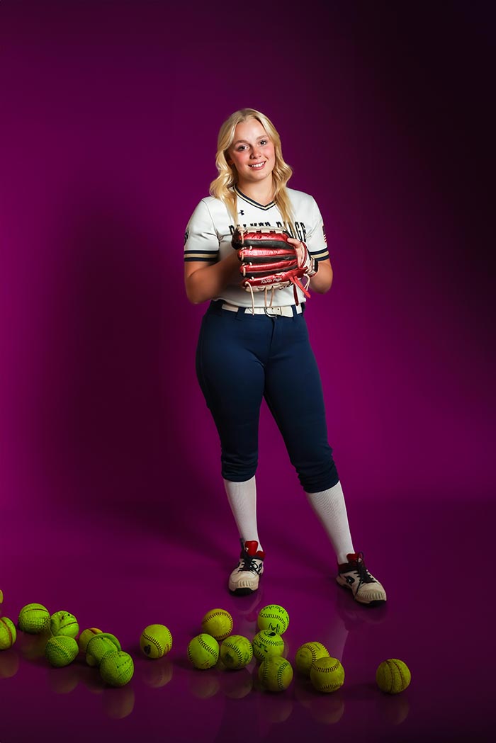 Vibrant studio sports portrait of female softball player in uniform holding glove with scattered softballs on purple backdrop, dynamic athlete photography in Colorado Springs showcasing strength and personality.