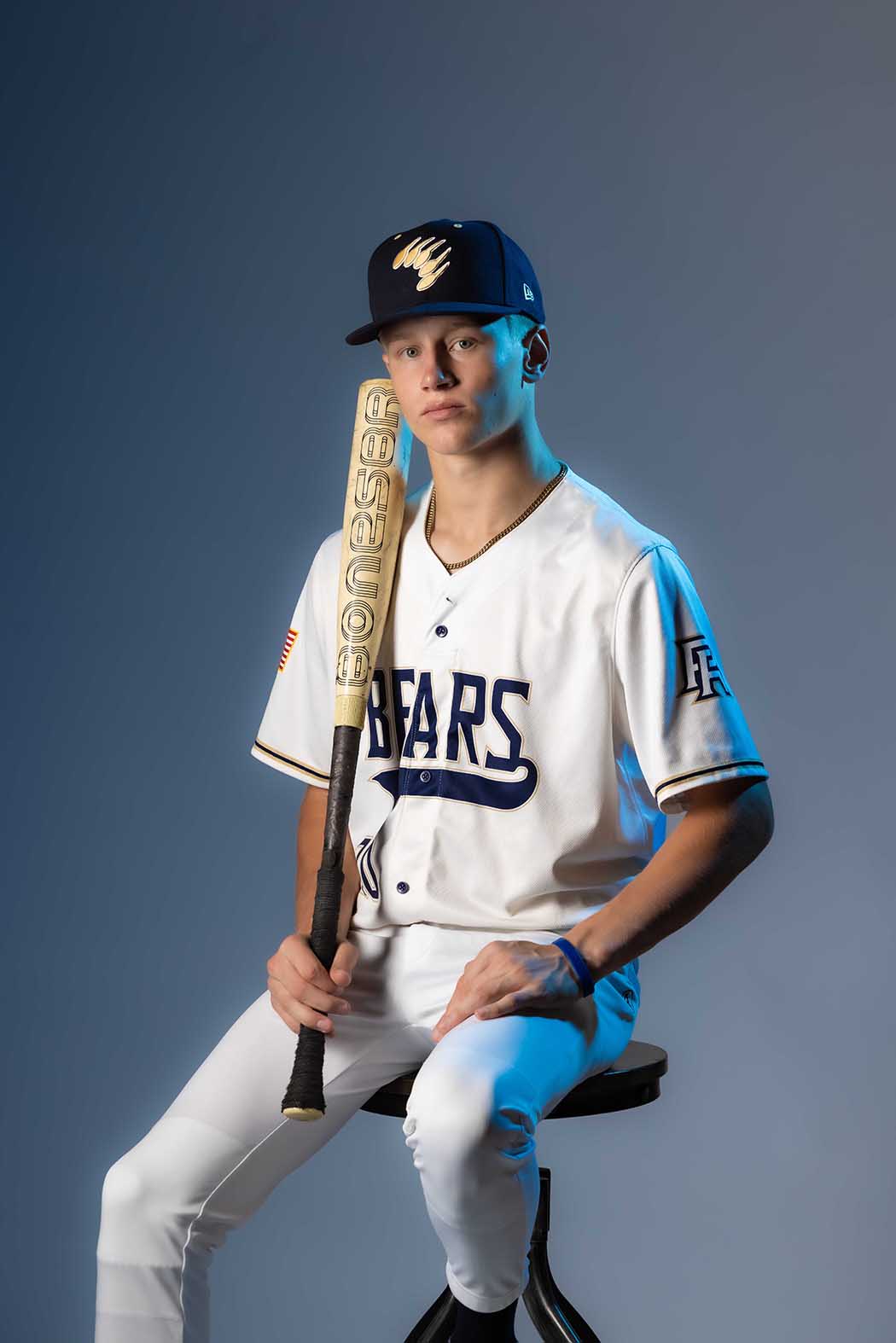 Studio sports portrait of high school baseball player in uniform holding a bat, seated against a gradient blue background, professional athlete photography in Colorado Springs showcasing team pride and focus.