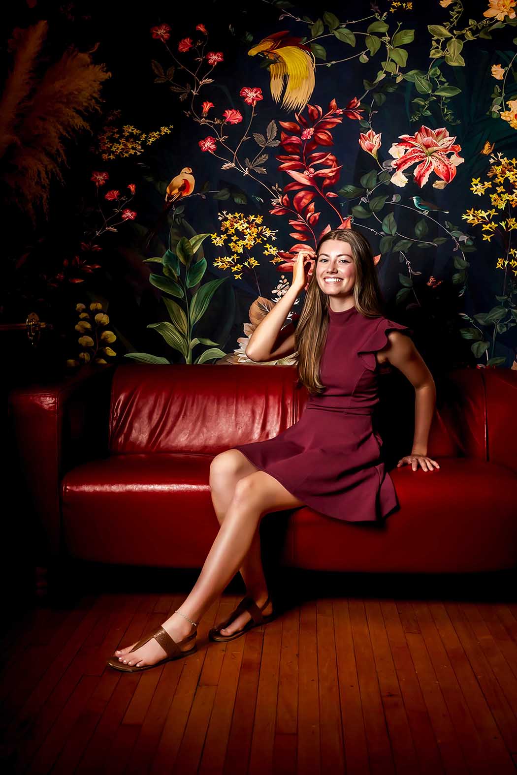 Stylish portrait of young woman in burgundy dress seated on red leather sofa with bold floral wallpaper backdrop, artistic lifestyle photography in Colorado Springs with vibrant colors and elegant composition.