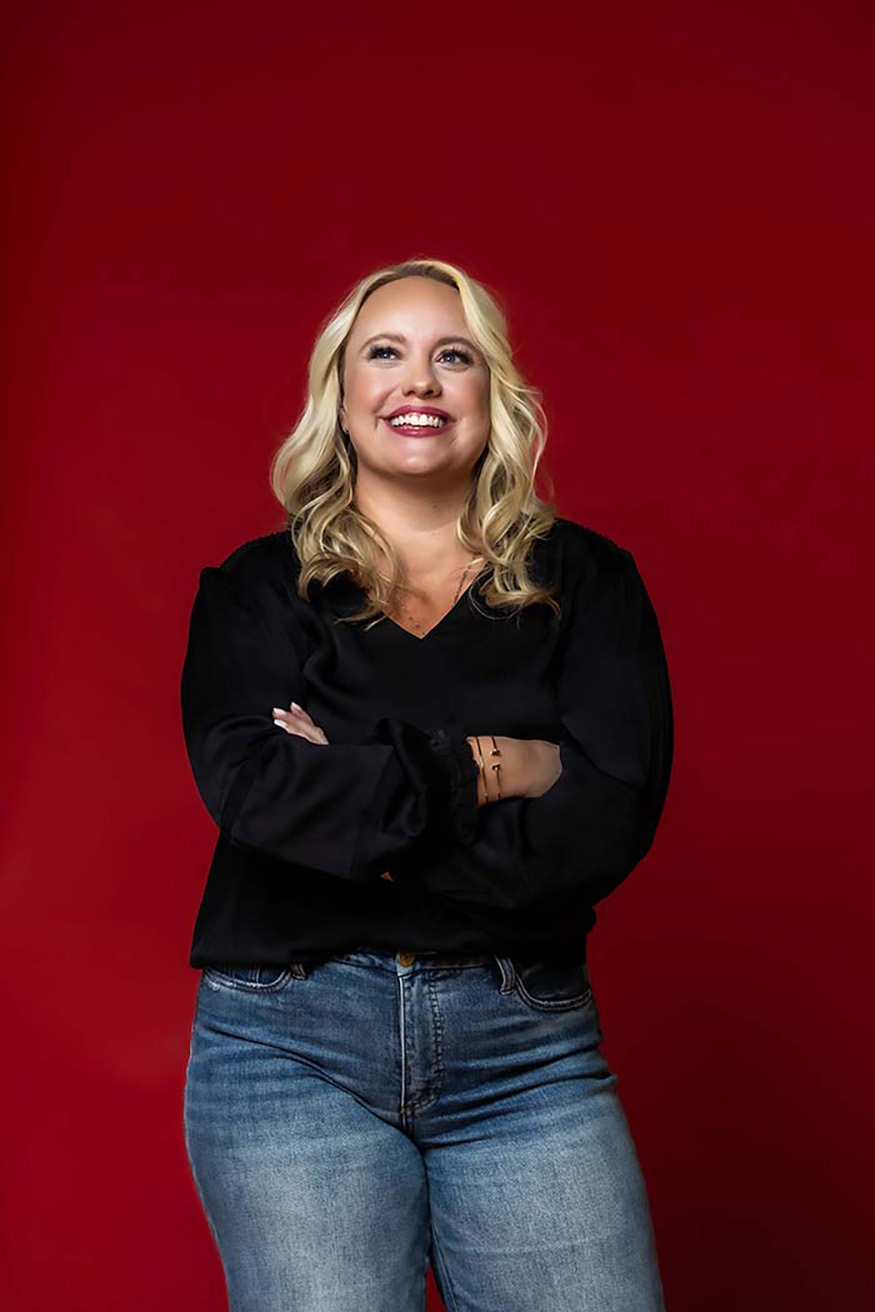 Studio branding portrait of smiling woman in black blouse and jeans standing confidently against a bold red background, modern lifestyle photography in Colorado Springs capturing authenticity and approachability.