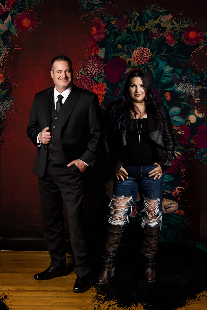 Stylized studio portrait of a professional man in a black suit and a confident woman in a leather jacket and distressed jeans, standing against a dark floral artistic backdrop, modern branding photography in Colorado Springs with contrasting fashion and personality.