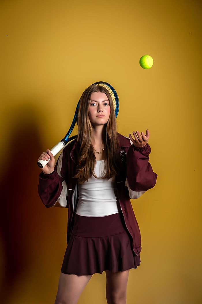 Studio portrait of confident teenage tennis player in maroon uniform tossing a neon tennis ball while holding a racquet, bold athletic branding photography in Colorado Springs with yellow background and strong attitude.