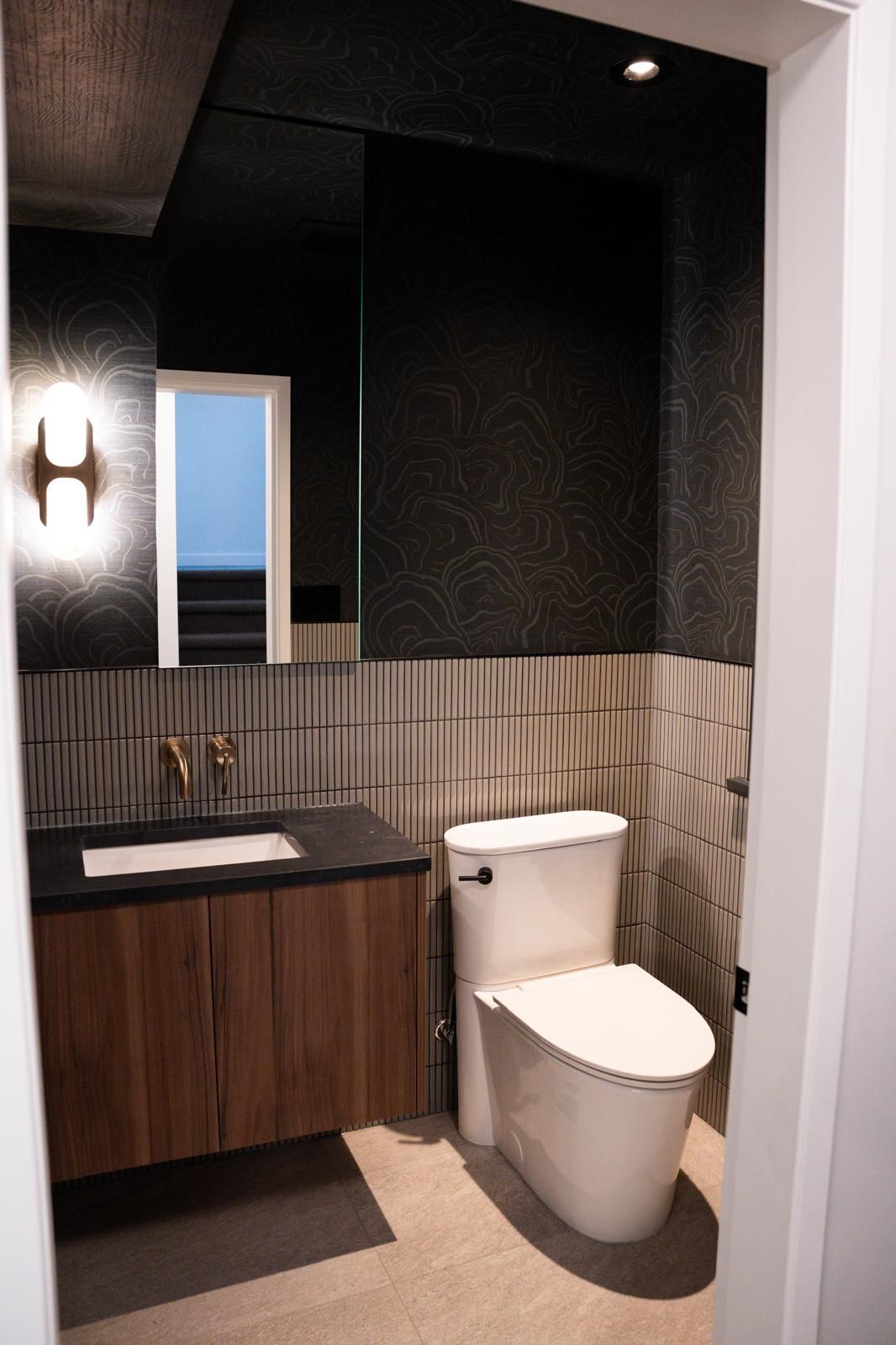 Renovated powder room with walnut floating vanity, dark tile walls, brass wall sconce and wall-hung toilet