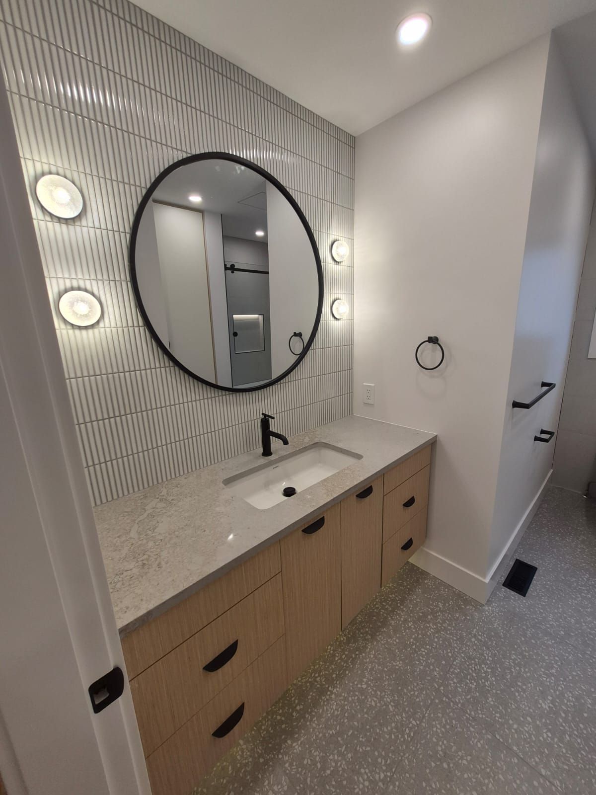 Bathroom renovation with round black mirror, white tile wall and light wood vanity