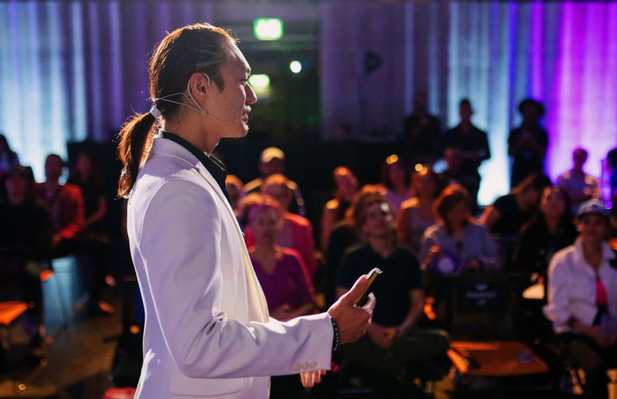 A person in a white suit speaking at a conference, addressing a seated audience.