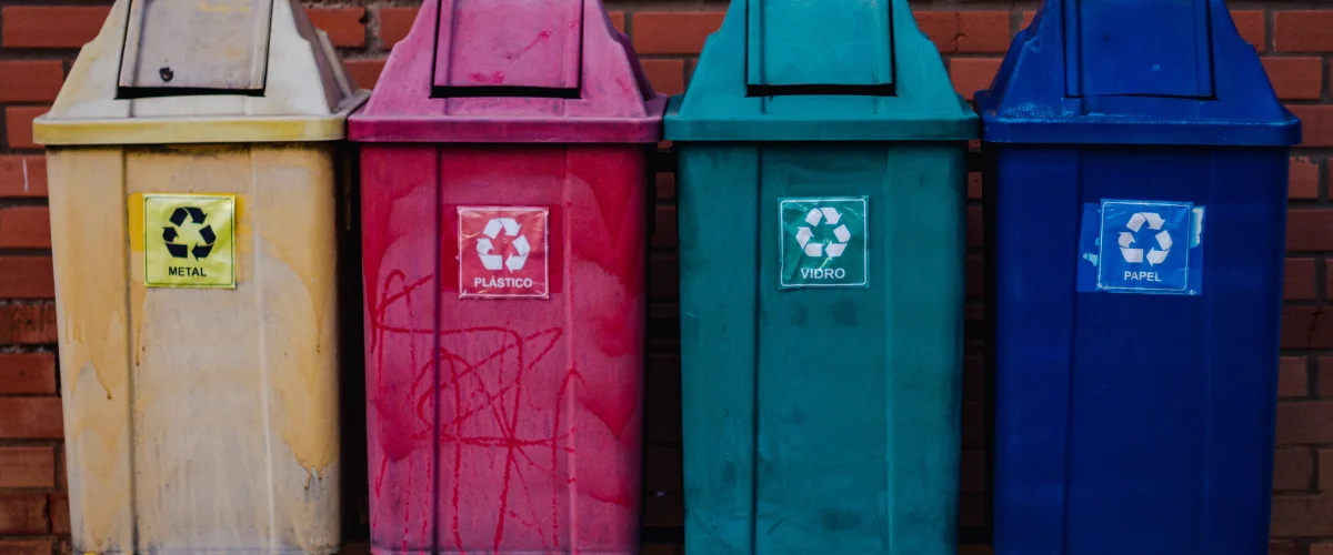 Four colorful recycling bins, yellow for metal, pink for plastic, teal for glass, and blue for paper, stand against a brick wall.