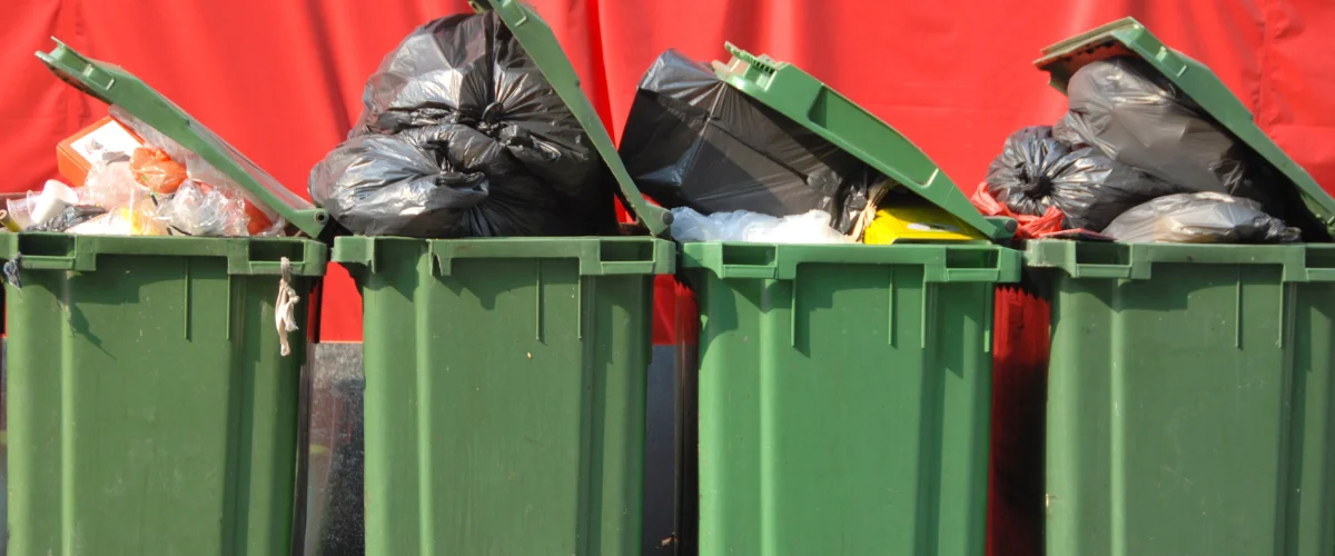 Four green waste bins overflowing with black and clear plastic bags of trash, set against a bright red background.