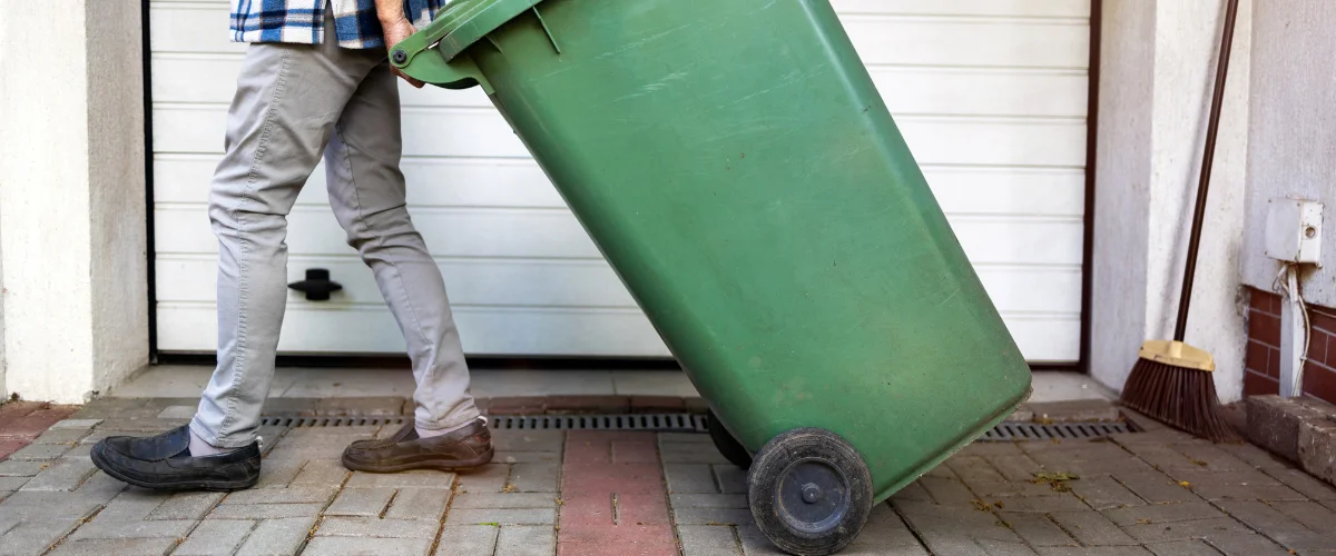 A man in gray pants and a plaid shirt pulls a large green wheeled trash bin across a paved driveway.