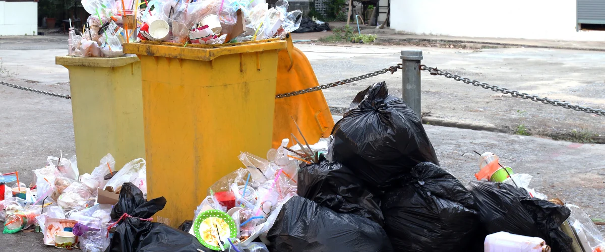 Two yellow street bins overflowing with plastic waste, surrounded by black trash bags and loose litter on concrete.