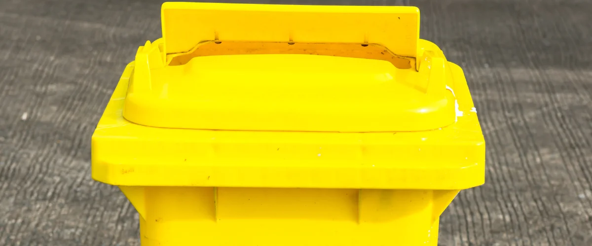 A bright yellow overflowing trash can with an open lid next to a black bin and bag, against a green wall.