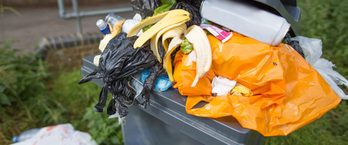 An overflowing outdoor rubbish bin filled with various waste items, including banana peels, plastic bags, and a soda can.