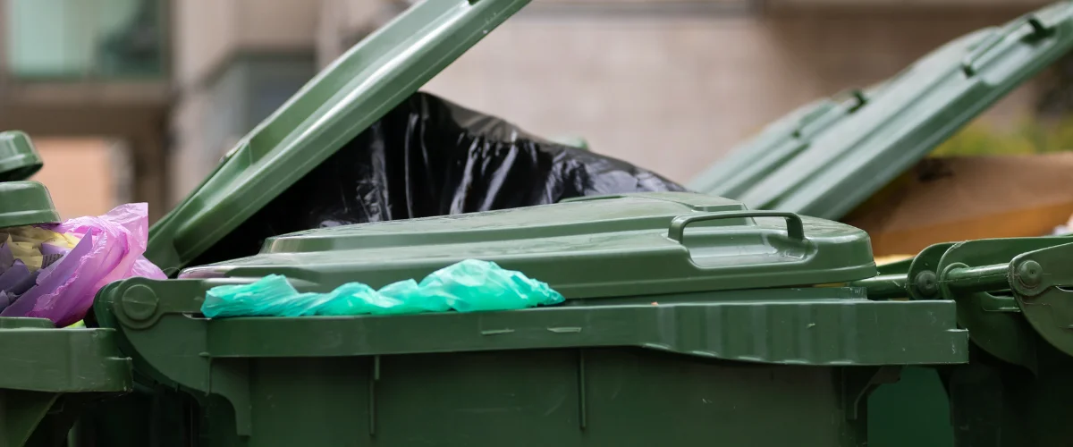 Multiple overflowing green dumpsters with lids ajar, revealing garbage bags and plastic waste.
