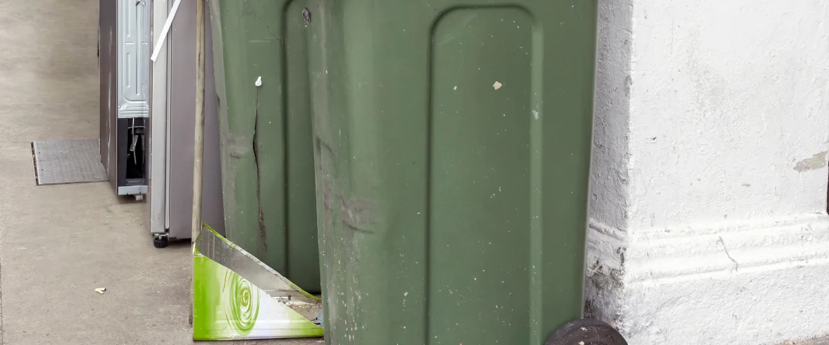 Two large green recycling bins and discarded refrigerators sit along a grey concrete wall with a white base.
