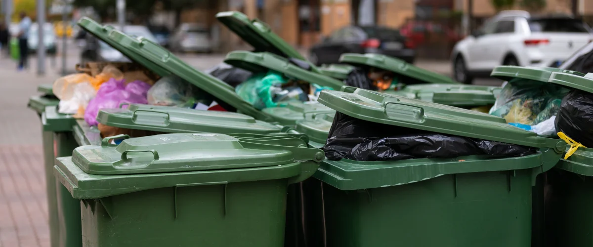 A row of overflowing green dumpsters with lids ajar, filled with colorful plastic bags of trash on a city street.