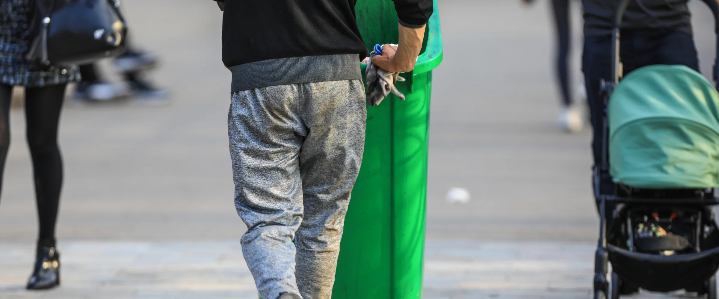 A person in grey sweatpants stands by a green bin, while a stroller and a person in black tights walk past.