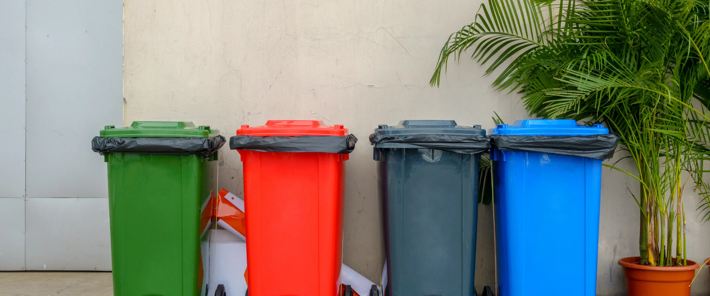 Four dark teal garbage bins with black liners and wooden blocks on top, lined up against a lush green fence. Four dark teal garbage bins with black liners and wooden blocks on top, lined up against a lush green fence.